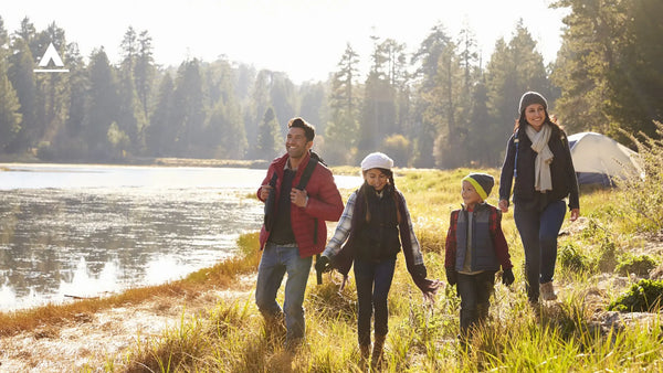 Photograph of family of 4 walking alongside river with hiking and camping gear towards campsite with tent, lit in bright sunny day with forest behind them and to the right of them.