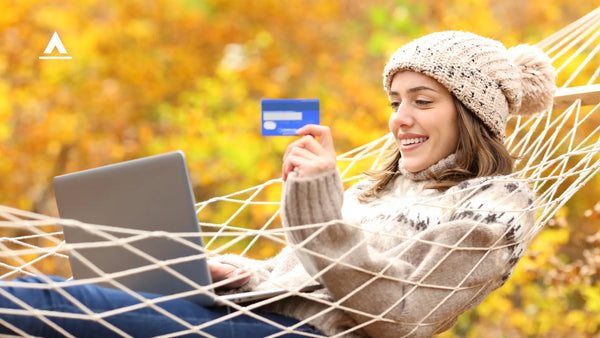 Woman sitting in hammock happily looking at debit card with autumn yellow trees in background.